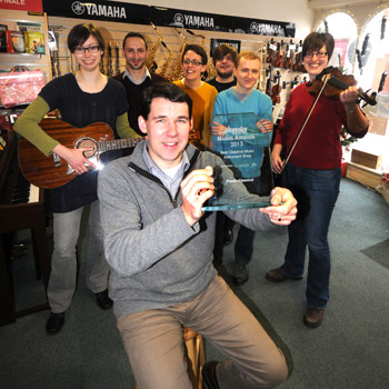 In the shop, Chris O'Reilly shows off the award, while behind him 6 members of staff display various instruments.